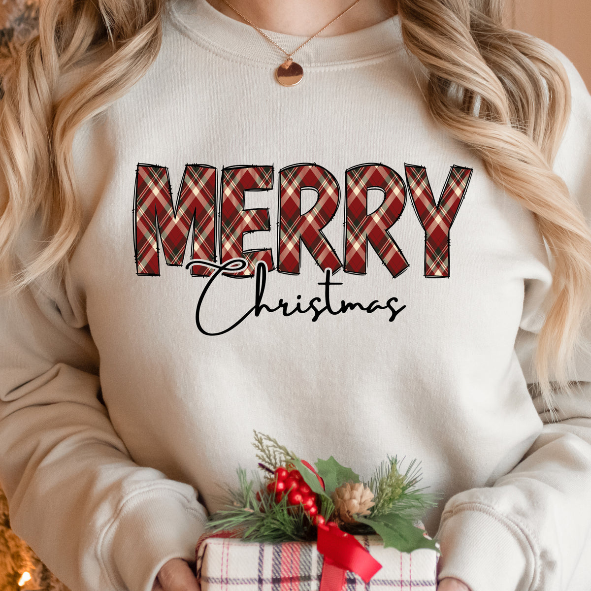 A woman wearing a sand Gildan sweatshirt with Merry in plaid and Christmas below that in script font.