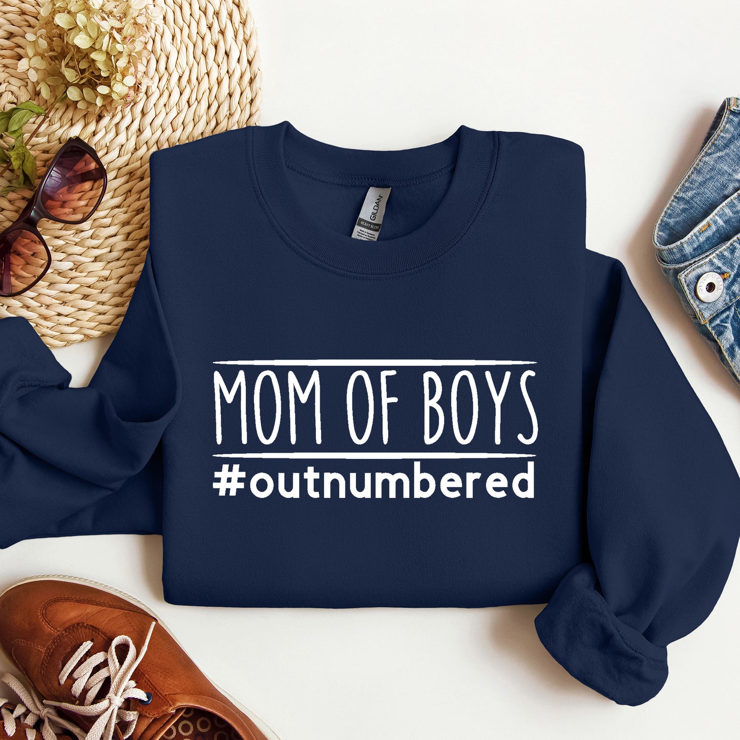 Navy blue sweatshirt with 'Mom of Boys #outnumbered' text on a white surface with accessories.