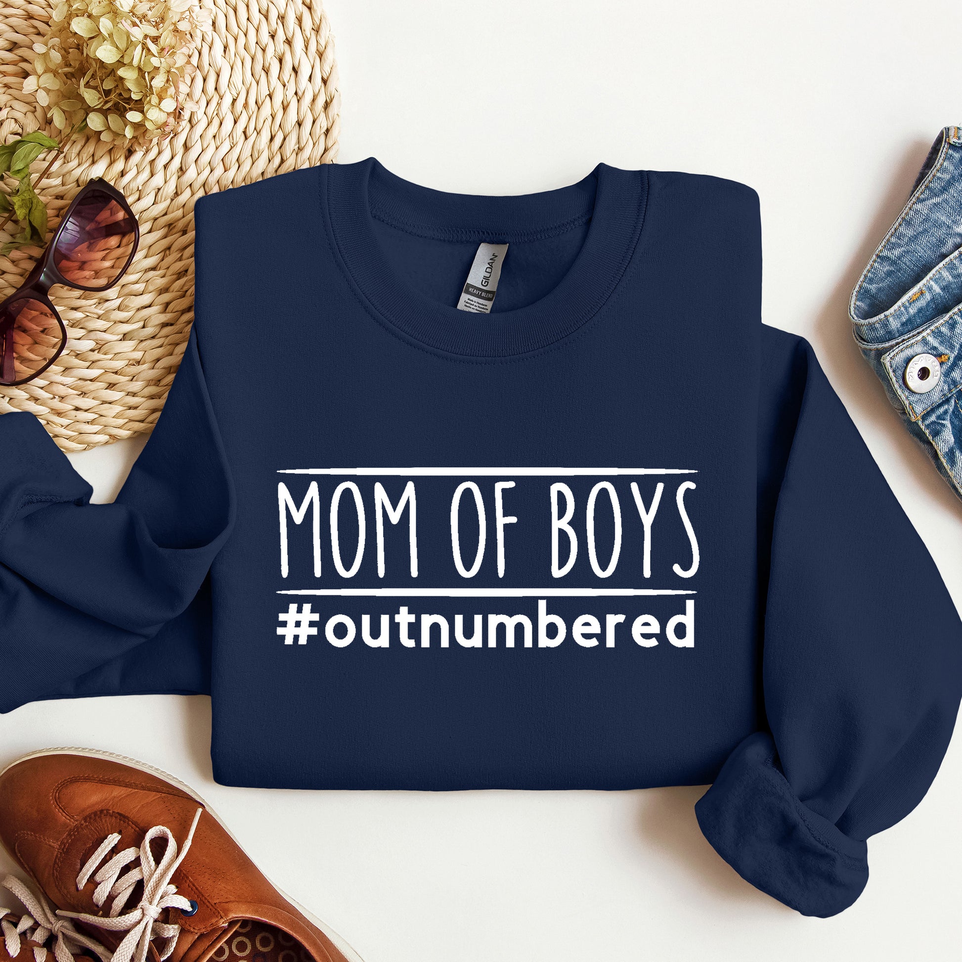 Navy blue sweatshirt with 'Mom of Boys #outnumbered' text on a white surface with accessories.