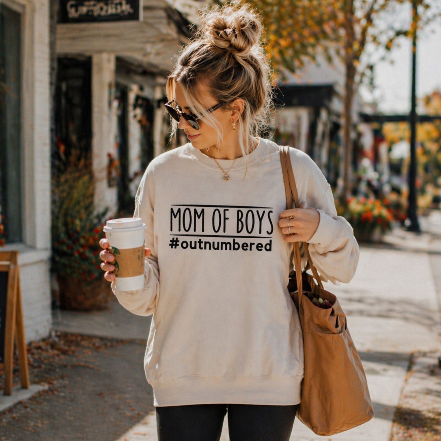 Woman wearing a sweatshirt with 'Mom of Boys #outnumbered' text, holding a coffee cup and a bag on a sidewalk.