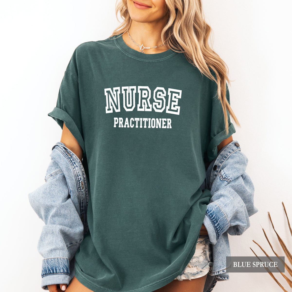 A woman wearing an oversized dark teal-green t-shirt with 'NURSE PRACTITIONER' printed in white block letters on the front, styled with rolled-up light-wash denim sleeves and denim shorts.