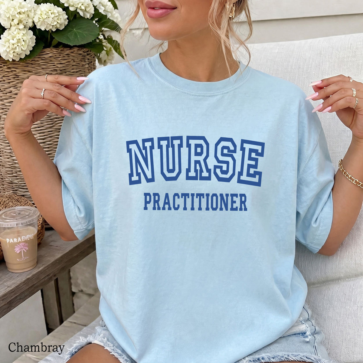 A woman wearing a light chambray blue oversized t-shirt with 'NURSE PRACTITIONER' printed in dark blue collegiate block letters across the chest, sitting on a white couch.