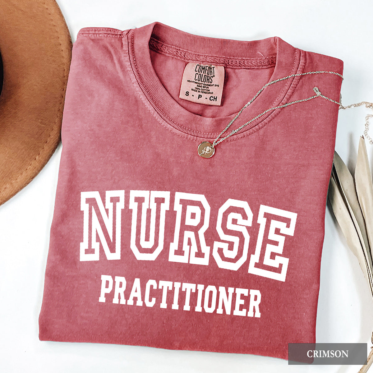 A folded dusty rose/terracotta t-shirt with 'NURSE PRACTITIONER' printed in white varsity letters on the front. The shirt is styled with a silver necklace and a brown hat on a white background.