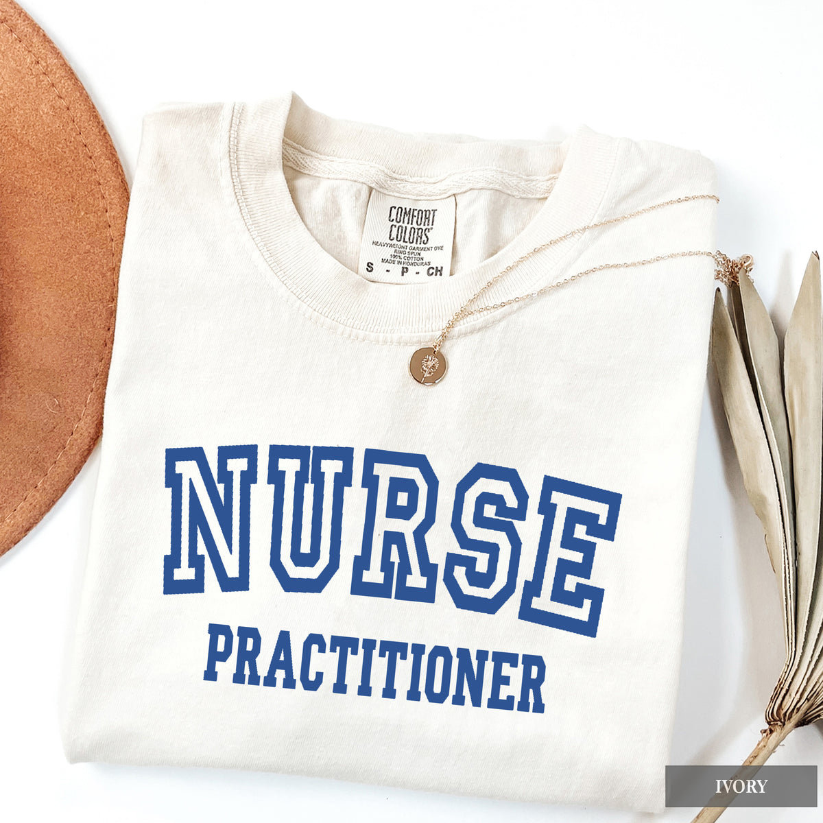 A folded ivory crewneck t-shirt with 'NURSE PRACTITIONER' printed in blue varsity lettering, styled with a gold pendant necklace and dried leaves on a white background.