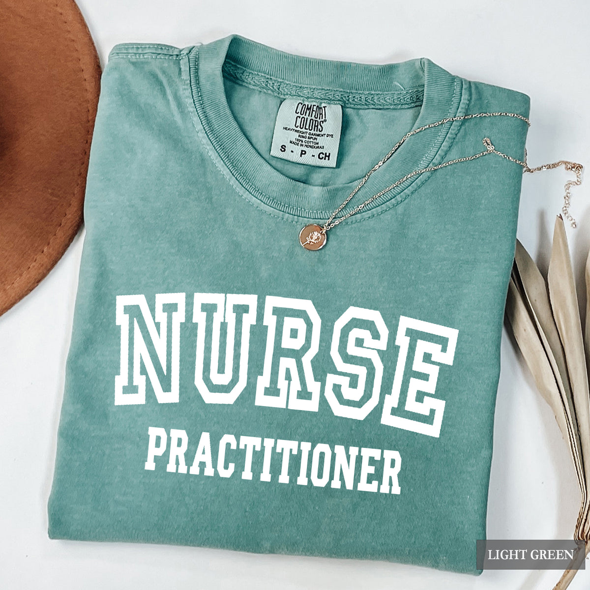 A folded light green crewneck t-shirt with 'NURSE PRACTITIONER' printed in white varsity lettering. The shirt is styled with a silver necklace and a brown leather hat on a white background.
