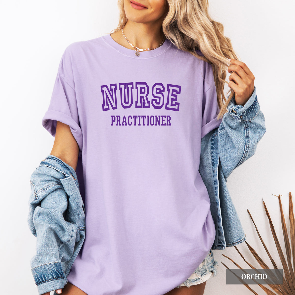 A woman wearing an oversized lavender-purple t-shirt with 'NURSE PRACTITIONER' printed in purple collegiate lettering. She has a light-wash denim jacket draped off her shoulders and is standing against a clean white background.