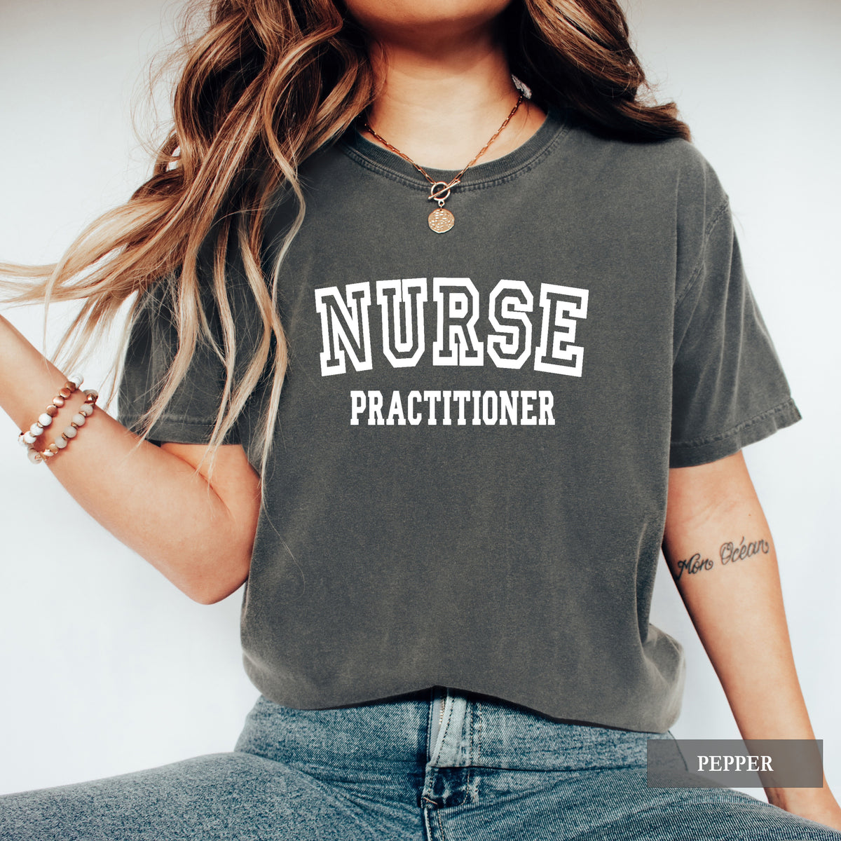 A woman wearing a faded charcoal gray t-shirt with 'NURSE PRACTITIONER' printed in white block letters on the front, paired with blue jeans. A 'PEPPER' color label is visible in the bottom right corner.
