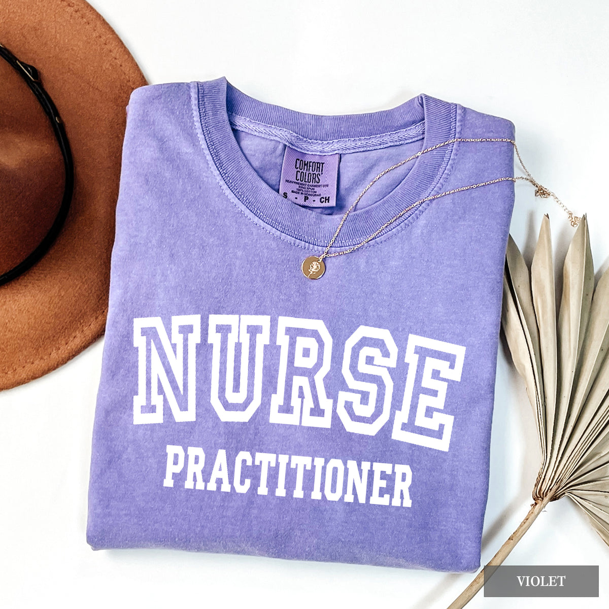 A folded lavender-purple t-shirt with 'NURSE PRACTITIONER' printed in white varsity lettering, styled in a flat lay with a brown hat, sunglasses, a gold necklace, and dried palm leaves on a white background.