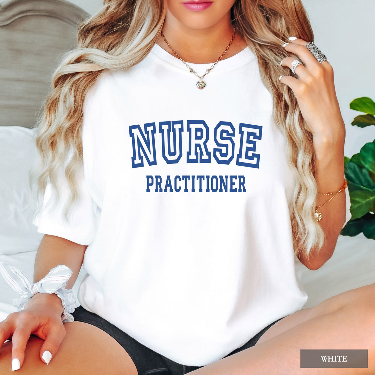 A woman wearing a white short-sleeve T-shirt with 'NURSE PRACTITIONER' printed in blue block letters across the chest. The photo is cropped from the lower face to the upper thighs, with a bed and plant in the background.