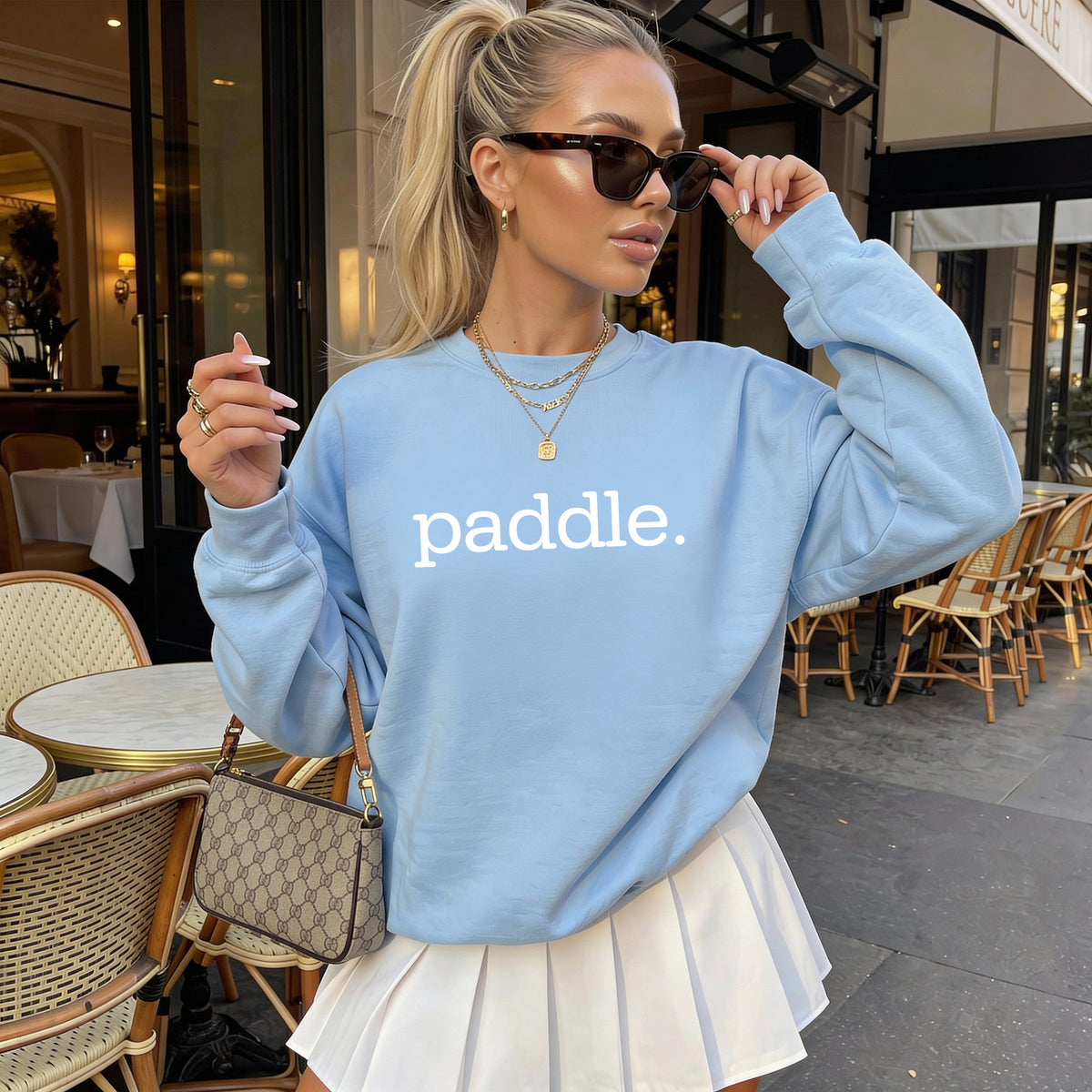 A woman wearing a light blue sweatshirt with the word 'paddle.' printed in white across the chest, styled with a white pleated skirt, sunglasses, and a monogram handbag in an outdoor cafe setting.