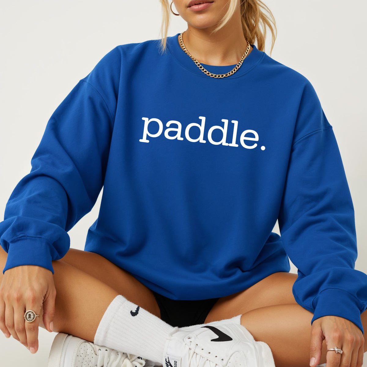 A woman wearing a royal blue crewneck sweatshirt with the white printed text 'paddle' across the chest, paired with white socks and white sneakers.