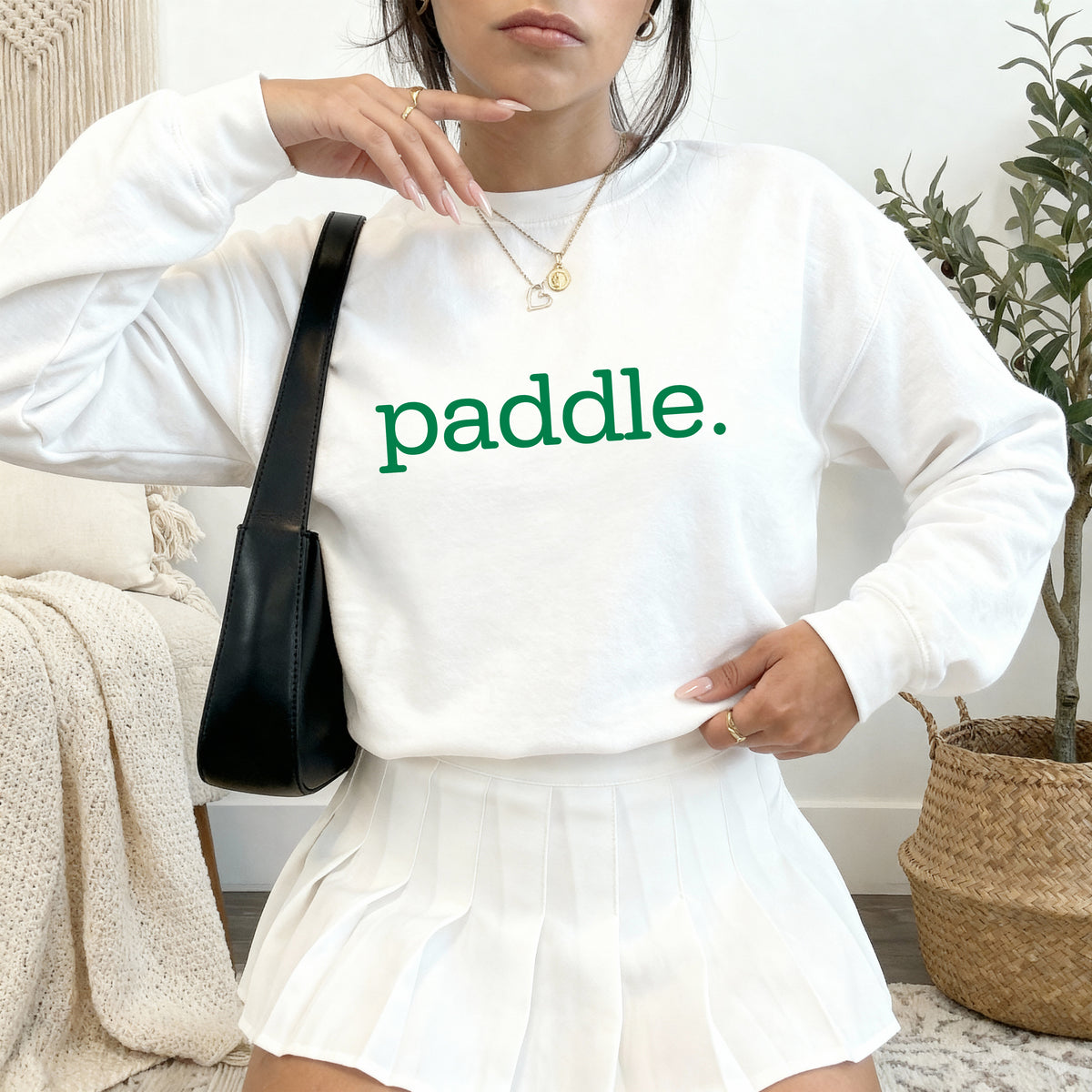 A woman wearing a white crewneck sweatshirt with the word 'paddle.' printed in green across the chest, paired with a white pleated skirt and carrying a black shoulder bag in a cozy indoor setting.