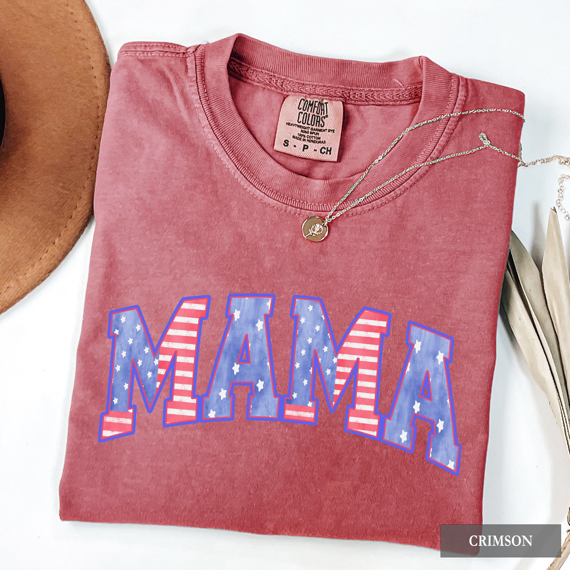 Folded red shirt with 'MAMA' in American flag design, next to a hat and necklace on a white background.