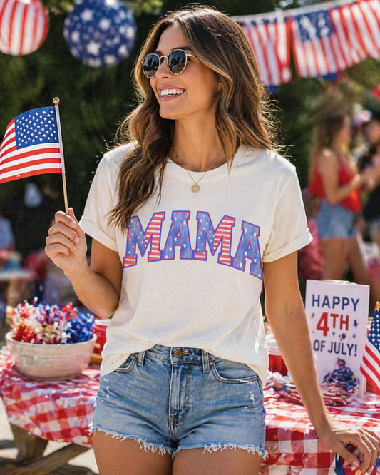Woman wearing a 'MAMA' t-shirt with American flag colors, holding a small American flag, standing in front of a 4th of July-themed table.