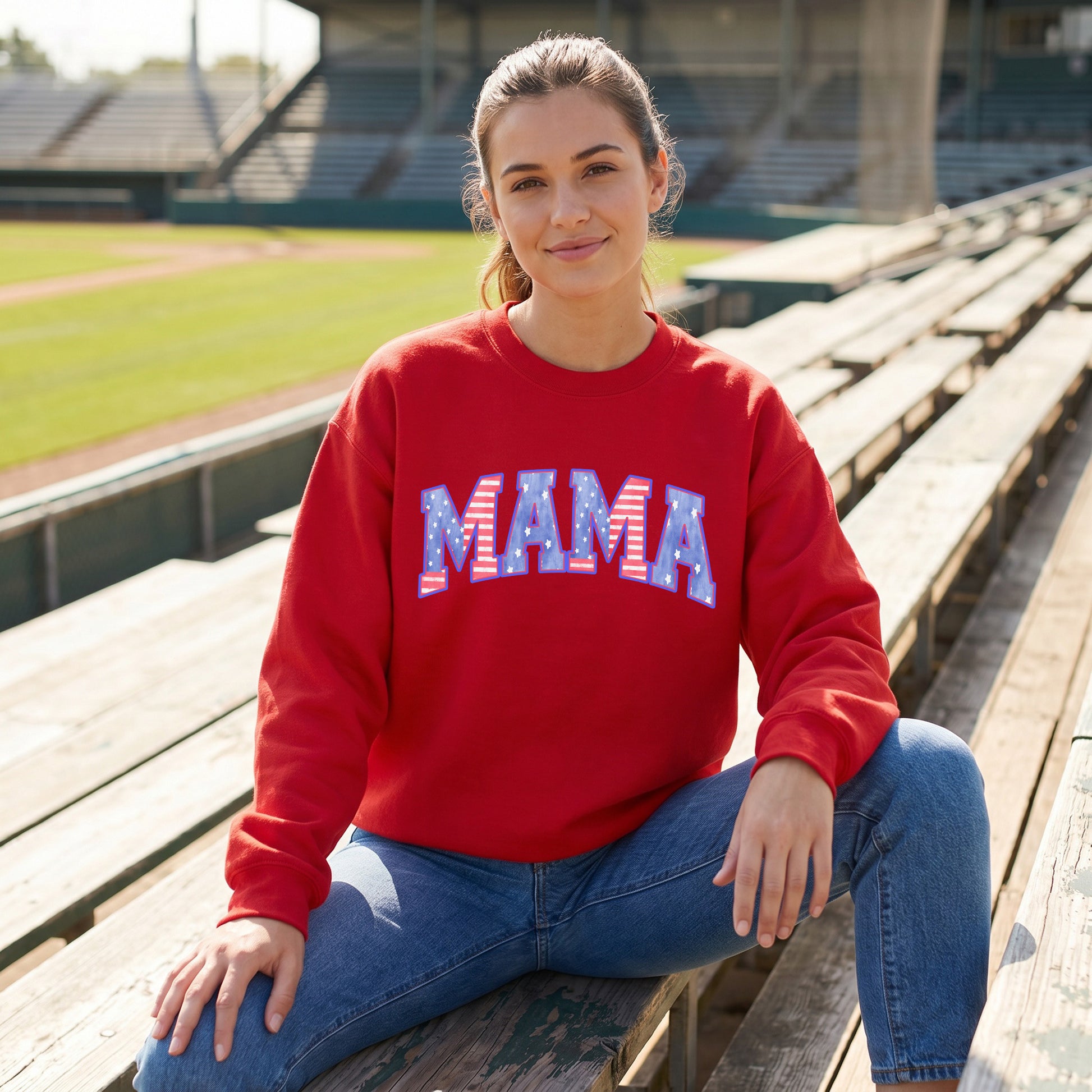 Woman wearing a red sweatshirt with 'MAMA' printed on it in a red white and blue American flag pattern, sitting on wooden bleachers at a stadium.