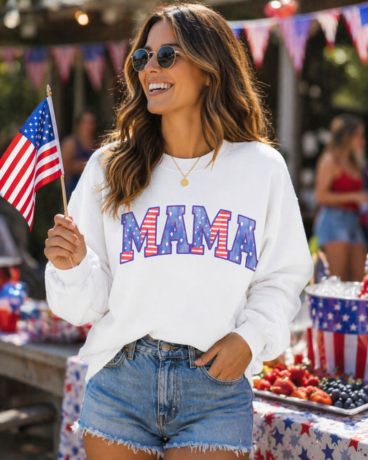 Woman wearing a white sweatshirt with 'MAMA' in colorful letters, holding an American flag, at a 4th of July event.