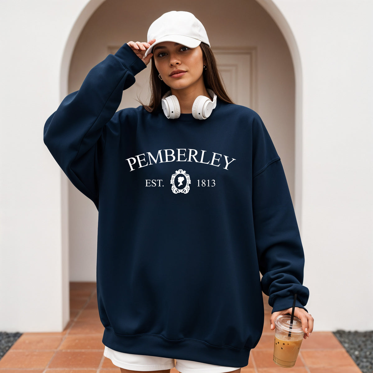 A woman wearing an oversized navy blue sweatshirt with 'PEMBERLEY EST. 1813' printed in white on the front, styled with a white baseball cap, white headphones around her neck, and holding an iced coffee in a tiled indoor-outdoor setting.