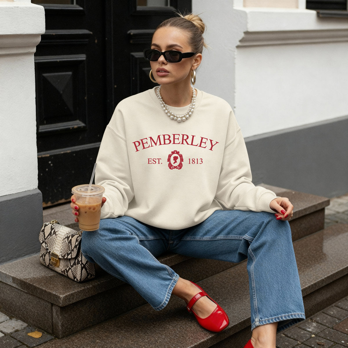 A woman sits on outdoor stone steps wearing an oversized cream sweatshirt with red 'PEMBERLEY EST. 1813' text and blue jeans, holding an iced coffee and wearing red flats.