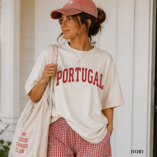 A woman wearing an oversized cream t-shirt with 'PORTUGAL' printed in red across the chest, paired with red-and-white gingham shorts, a dusty pink baseball cap, and a cream tote bag, standing in front of a white porch.