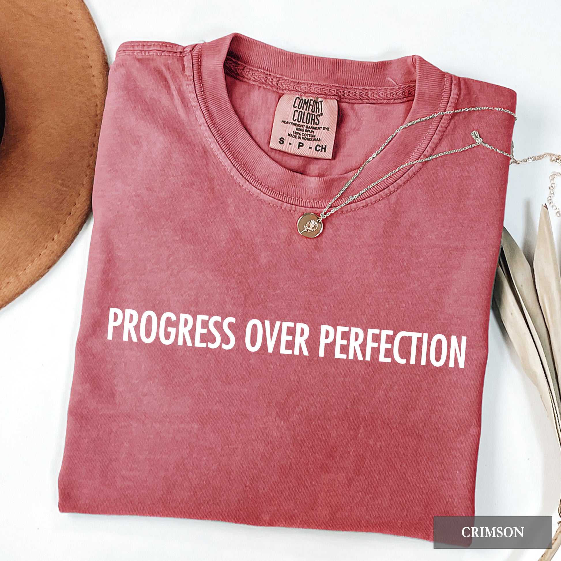 A folded crewneck t-shirt in a washed crimson color with the text 'PROGRESS OVER PERFECTION' printed in white across the chest. A silver necklace with a round pendant is draped over the collar.