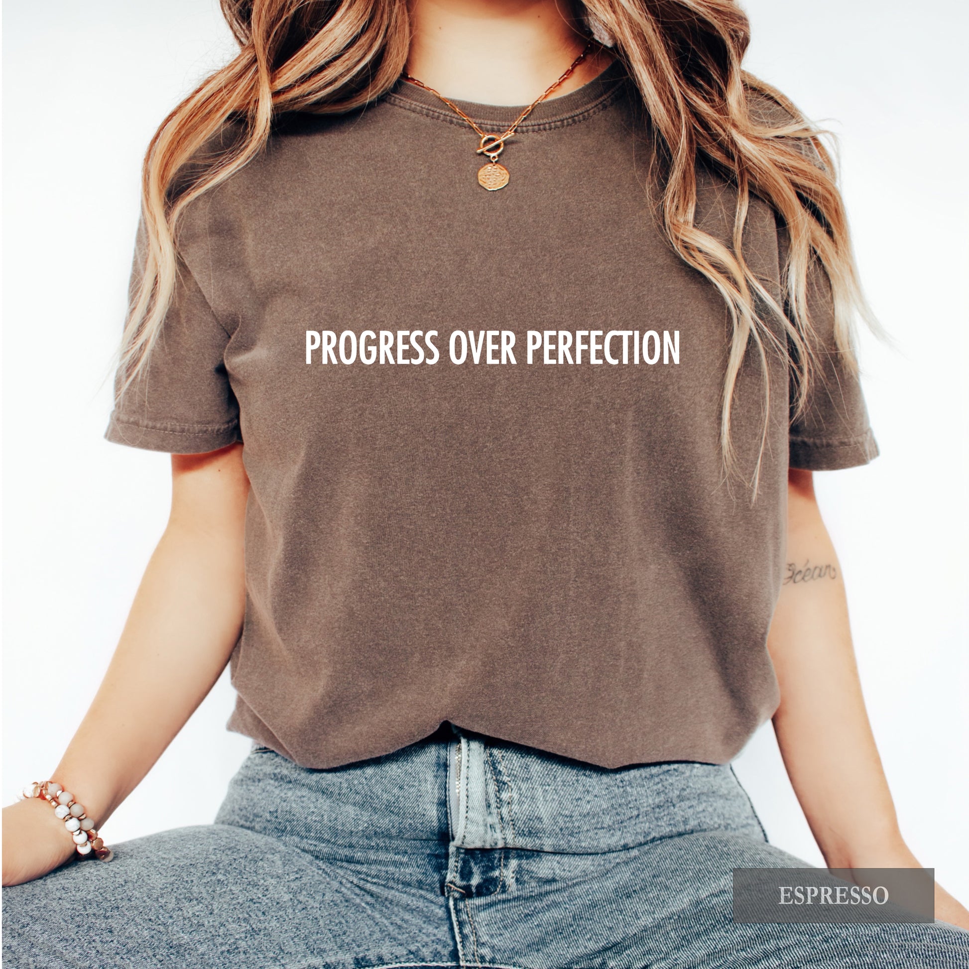 A woman wearing a cropped short-sleeve t-shirt in washed espresso brown with the text 'PROGRESS OVER PERFECTION' printed in white across the chest. She is also wearing blue jeans and a gold necklace, set against a plain white background.