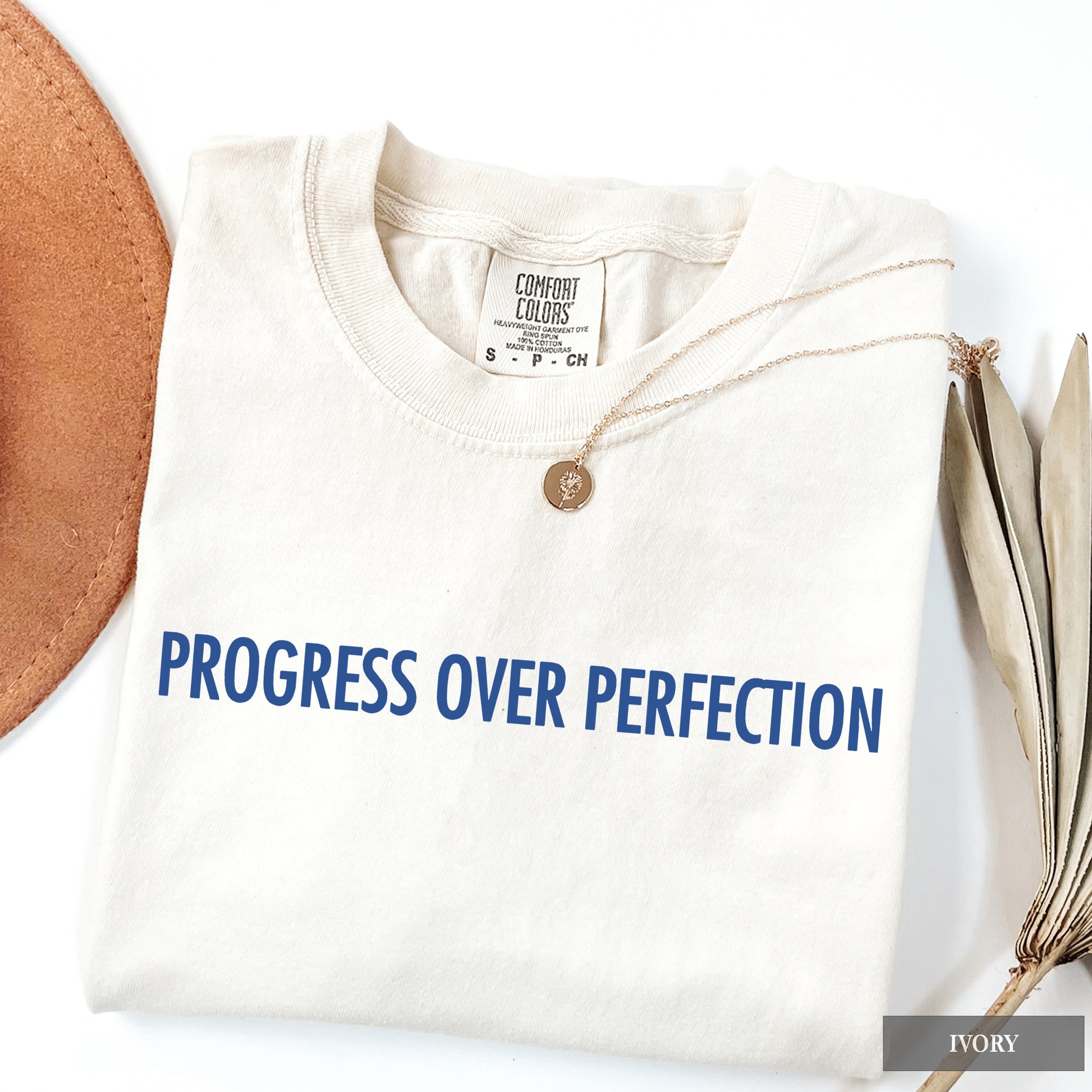 A folded ivory crewneck t-shirt with the phrase 'PROGRESS OVER PERFECTION' printed in blue across the chest. The shirt is styled with a small gold pendant necklace and a brown leather circle on a white background.