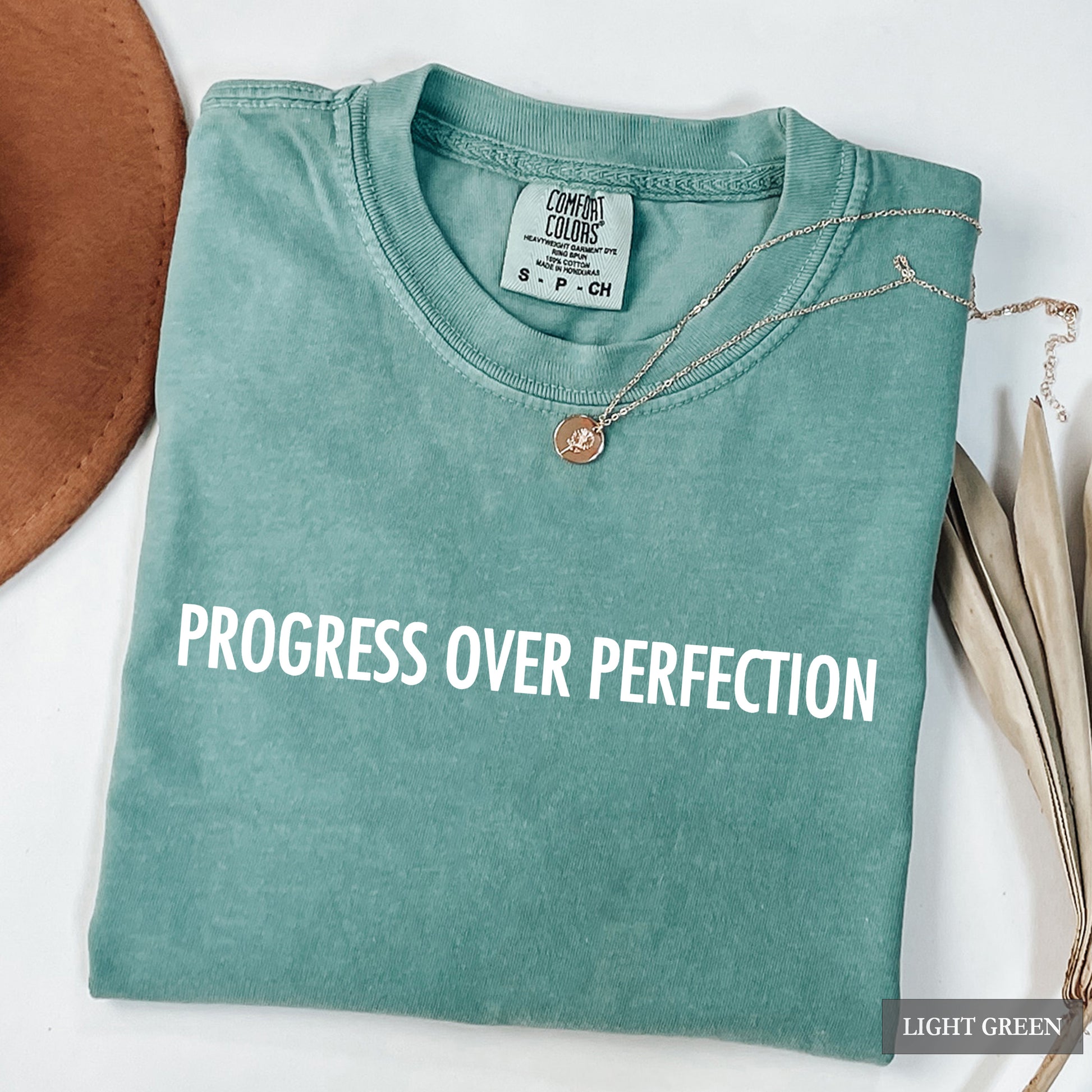 A folded light green crewneck t-shirt with the phrase 'PROGRESS OVER PERFECTION' printed in white across the chest. The shirt is styled with a delicate silver necklace and a brown leather hat edge on a white background.