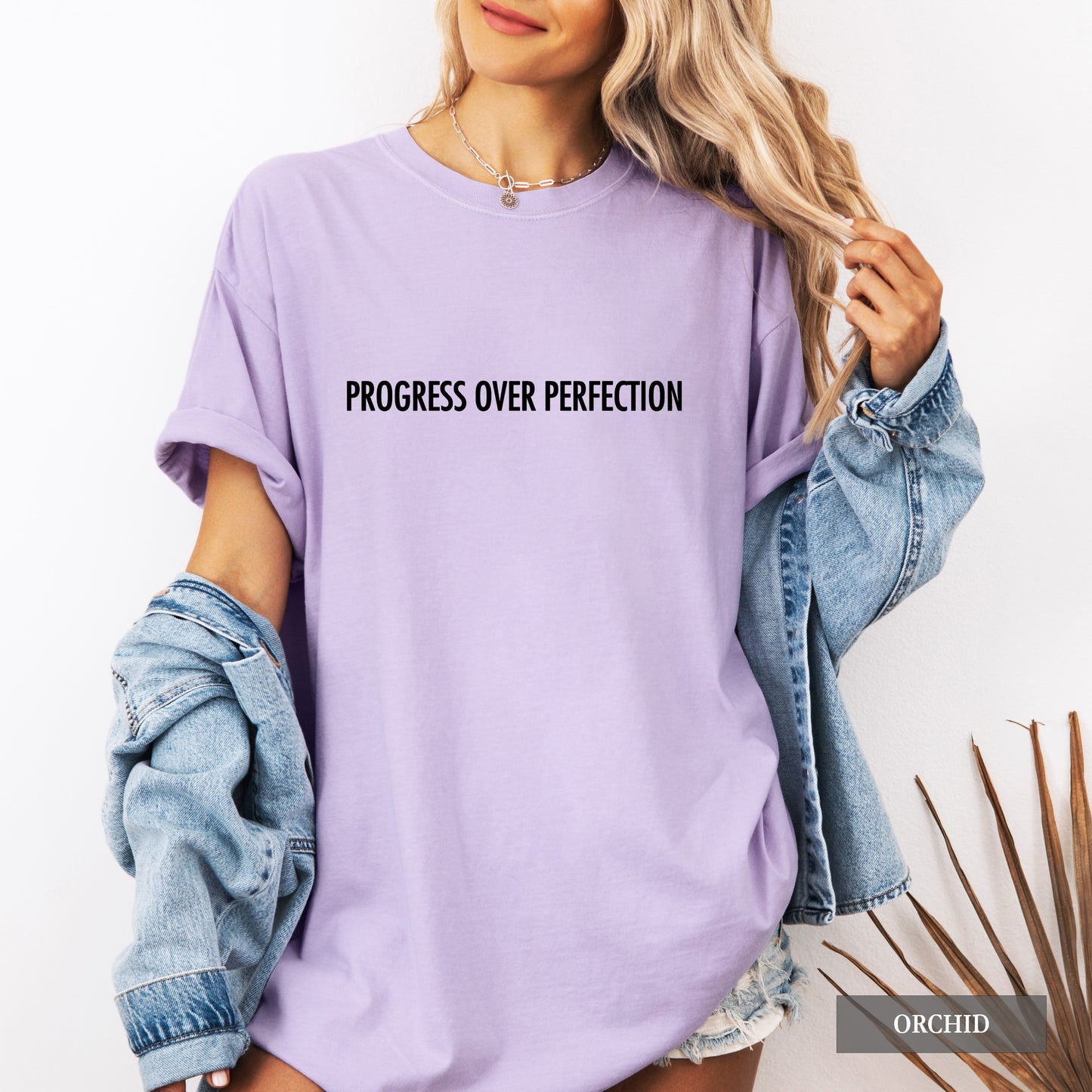 A woman wearing an oversized lavender-purple t-shirt with the text 'PROGRESS OVER PERFECTION' printed in black across the chest. She has a light-wash denim jacket draped off her shoulders and is posed against a clean white background.
