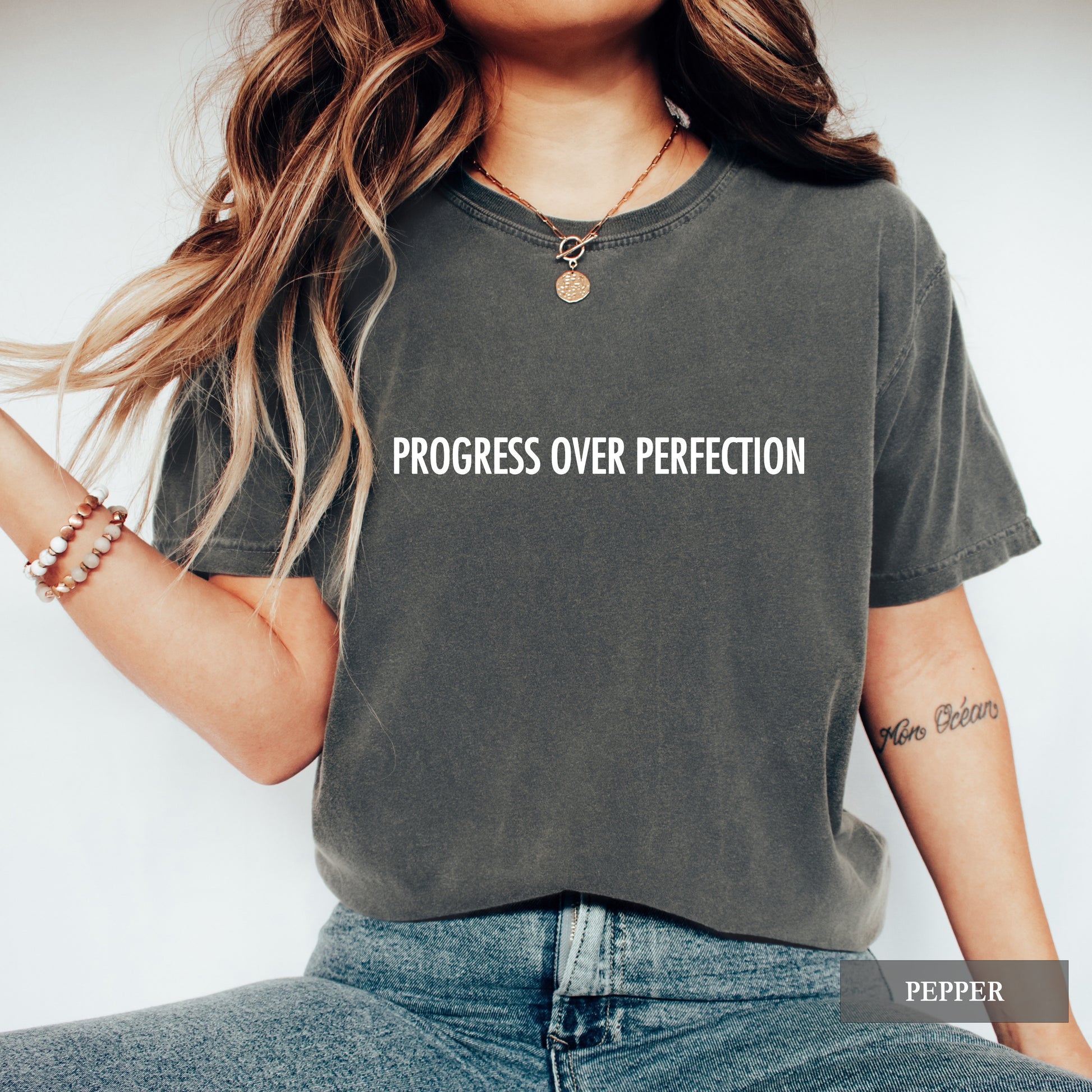 A woman wearing a faded charcoal gray t-shirt with the text 'PROGRESS OVER PERFECTION' printed in white across the chest. The image is a lifestyle shot with a white background, featuring a 'PEPPER' color label overlay in the bottom right corner.