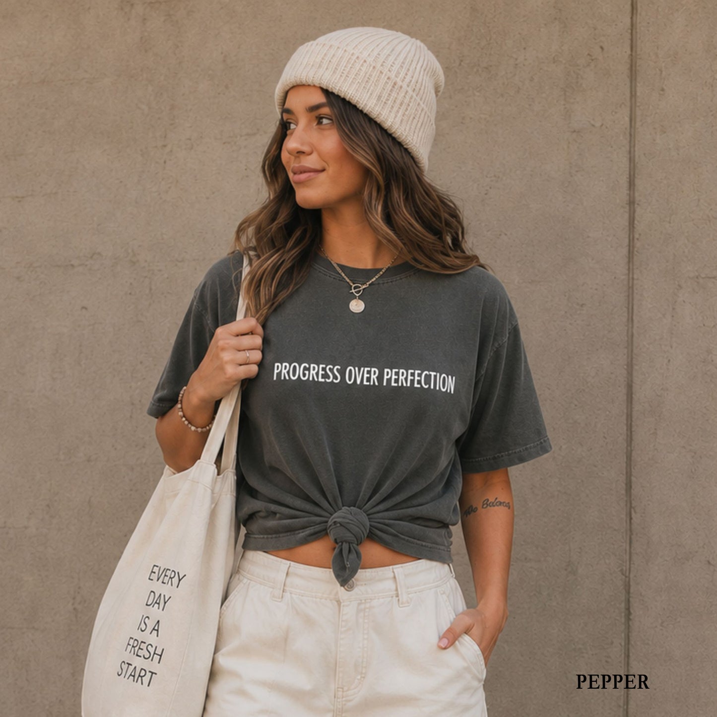 A woman wearing a dark gray graphic t-shirt with the words 'PROGRESS OVER PERFECTION' in white, tied at the waist. She is also wearing a cream knit beanie, white pants, and carrying a cream tote bag with black text.