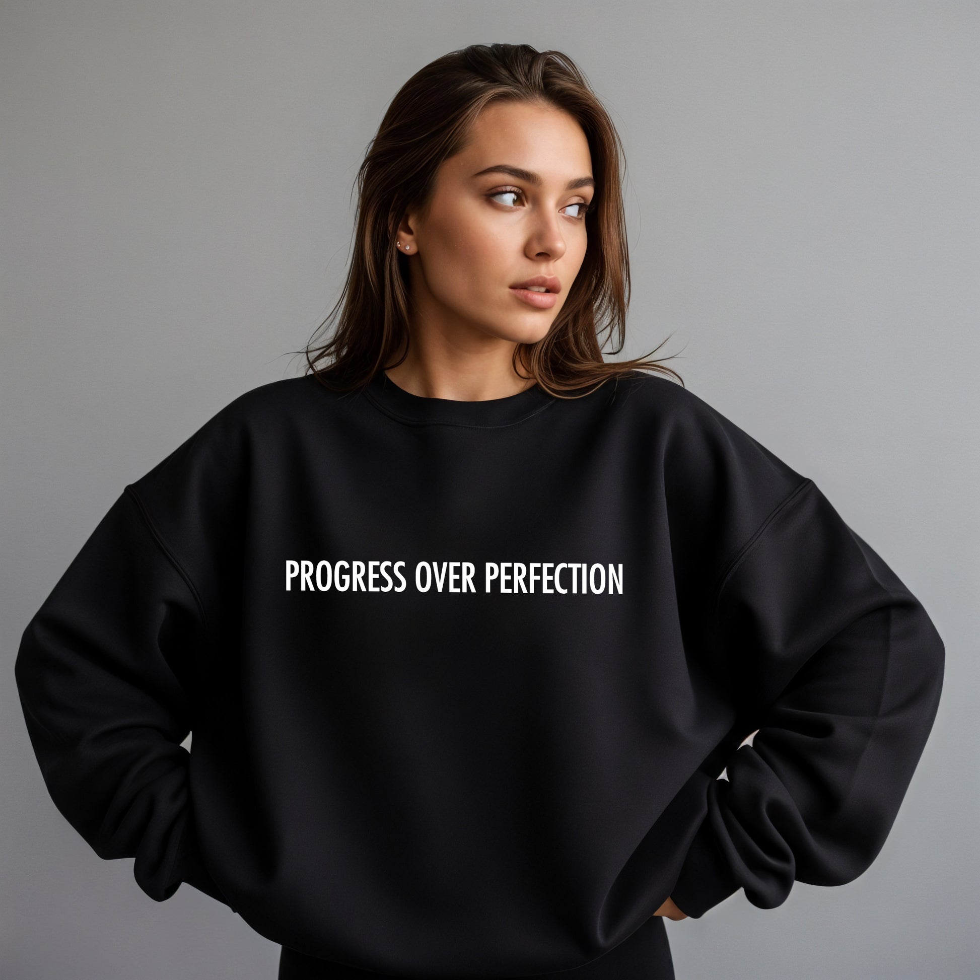 A woman wearing a black crewneck sweatshirt with the phrase 'PROGRESS OVER PERFECTION' printed in white capital letters across the chest, set against a plain gray studio background.