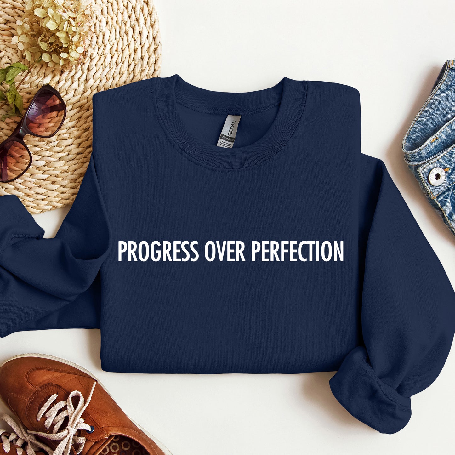 A navy blue crewneck sweatshirt with the phrase 'PROGRESS OVER PERFECTION' printed in white across the chest, styled in a flat lay with a straw hat, sunglasses, brown shoes, and denim jeans on a white background.