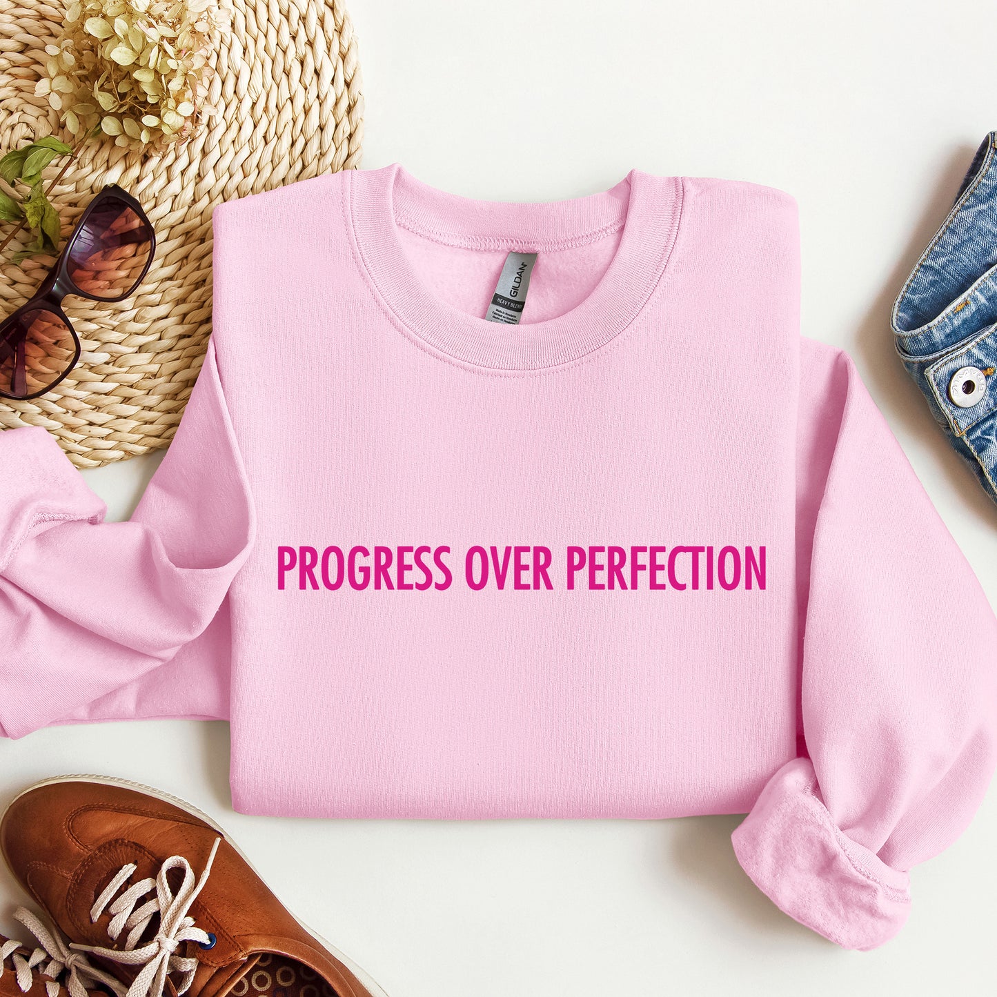 A folded light pink crewneck sweatshirt with the phrase 'PROGRESS OVER PERFECTION' printed in bright pink across the chest, styled in a flat lay with sunglasses, a woven hat, a brown shoe, and a denim item on a white background.