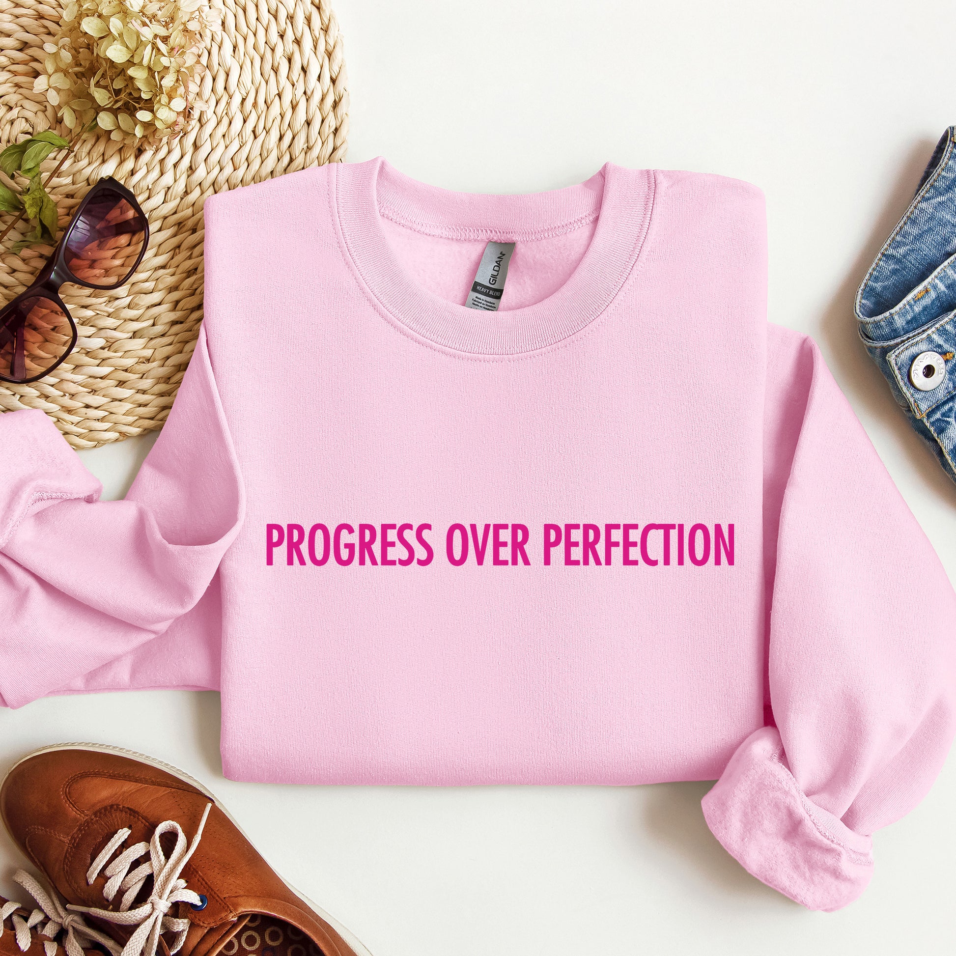 A folded light pink crewneck sweatshirt with the phrase 'PROGRESS OVER PERFECTION' printed in bright pink across the chest, styled in a flat lay with sunglasses, a woven hat, a brown shoe, and a denim item on a white background.