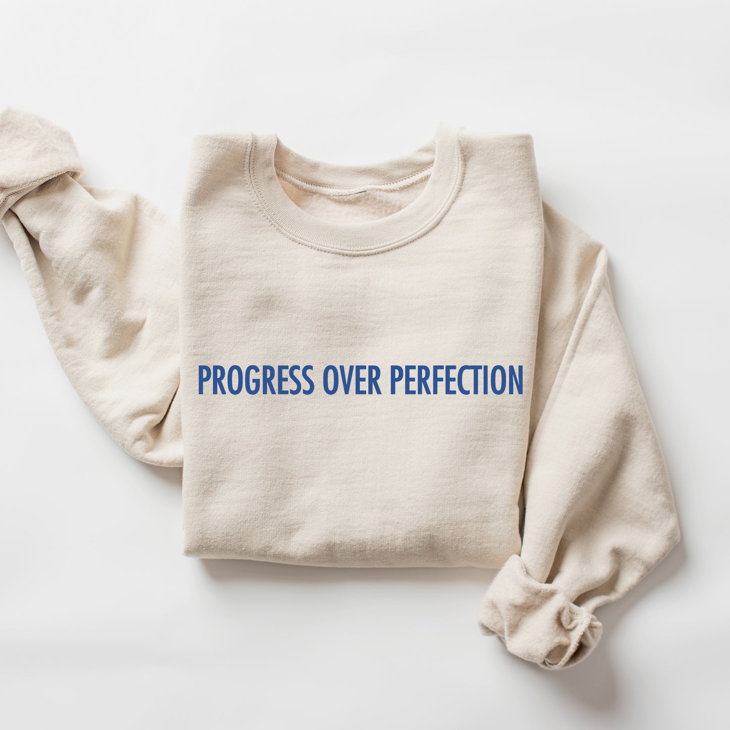A folded cream-colored crewneck sweatshirt with the phrase 'PROGRESS OVER PERFECTION' printed across the chest in blue capital letters, displayed on a clean white background.