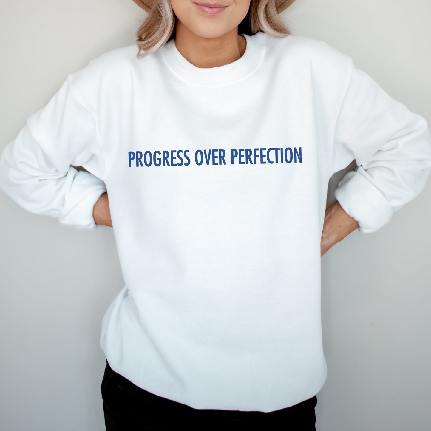A person wearing a white crewneck sweatshirt with the text 'PROGRESS OVER PERFECTION' printed in blue across the chest, posed against a light gray background.