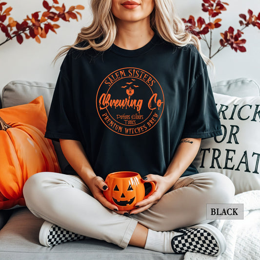 A woman sits cross-legged on a couch wearing a black graphic t-shirt with an orange circular 'Salem Sisters Brewing Co' Halloween design. She is holding an orange pumpkin mug, with a pumpkin and a 'Trick or Treat' pillow in the background.