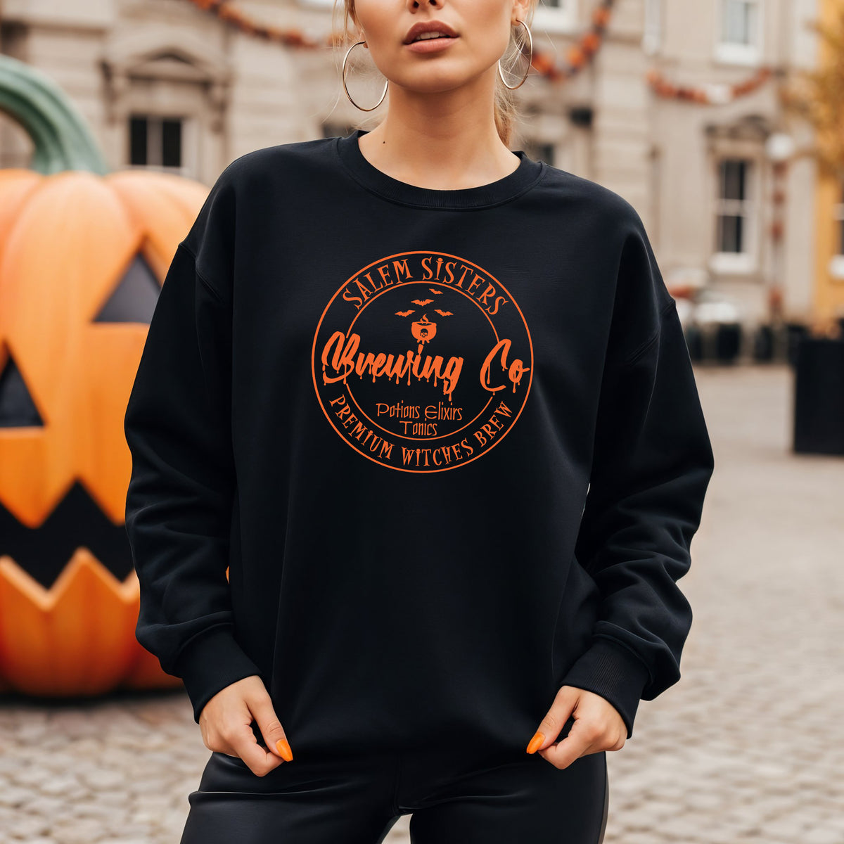 A woman wearing a black crewneck sweatshirt with a circular orange graphic that reads 'Salem Sisters Brewing Co' and 'Premium Witches Wear'. She is standing outdoors in front of a carved pumpkin and a blurred building background.
