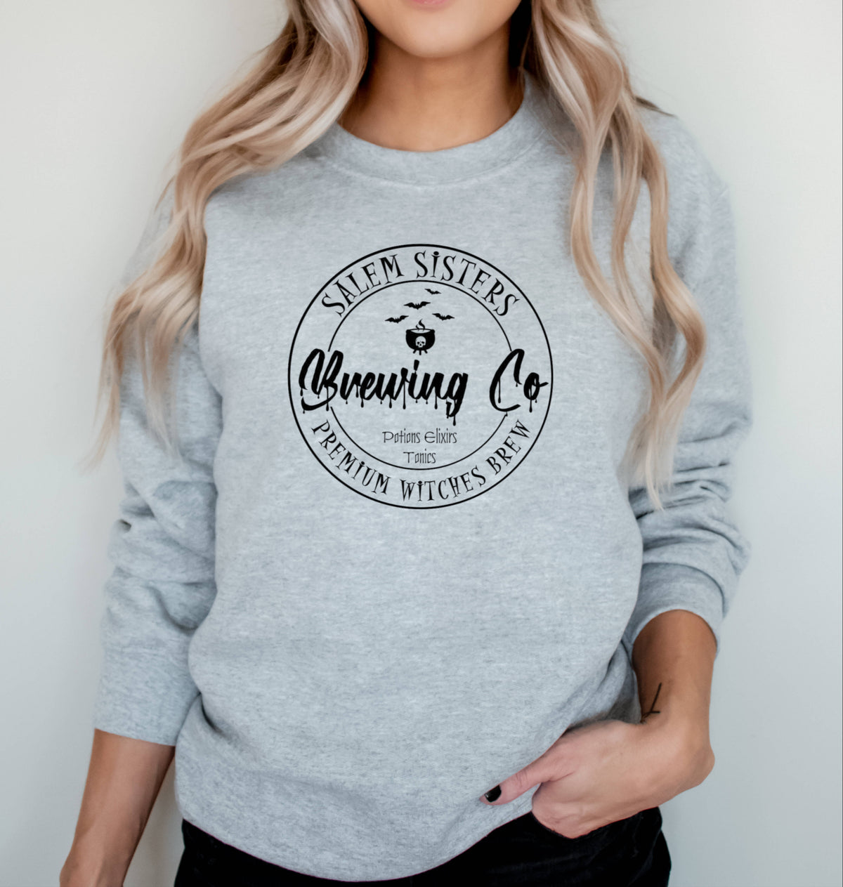 A woman wearing a light heather gray crewneck sweatshirt with a black circular graphic that reads 'Salem Sisters Brewing Co' and 'Premium Witches Brew'.