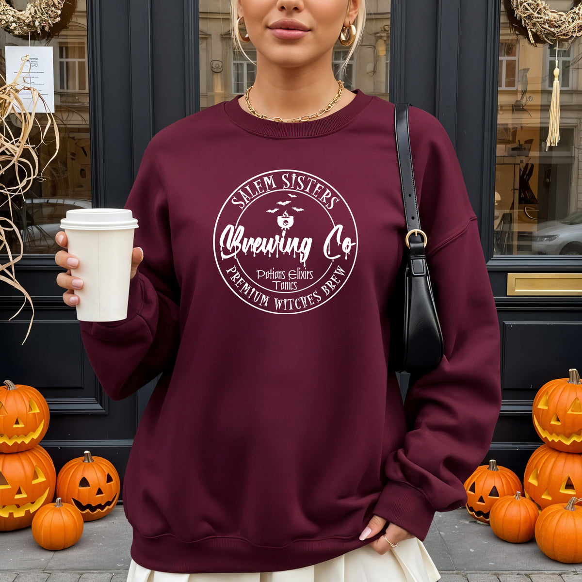A woman wearing a maroon crewneck sweatshirt with a white circular graphic that reads 'Salem Sisters Brewing Co' and 'Premium Witches Brew'. She is holding a white coffee cup in front of a dark doorway decorated with jack-o'-lanterns.
