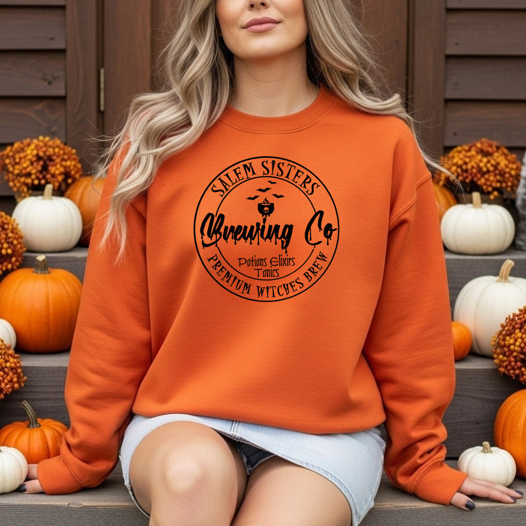 A woman wearing an orange crewneck sweatshirt with a black 'Salem Sisters Brewing Co' pumpkin graphic, posed on outdoor steps surrounded by pumpkins and fall decorations.