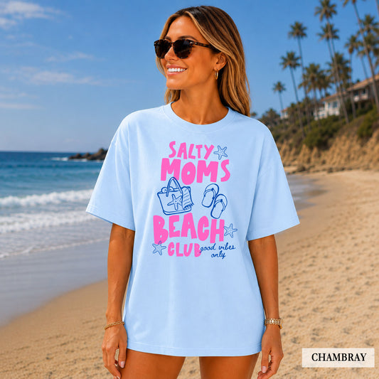 Woman wearing a light blue t-shirt with 'Salty Moms Beach Club' text on a beach.