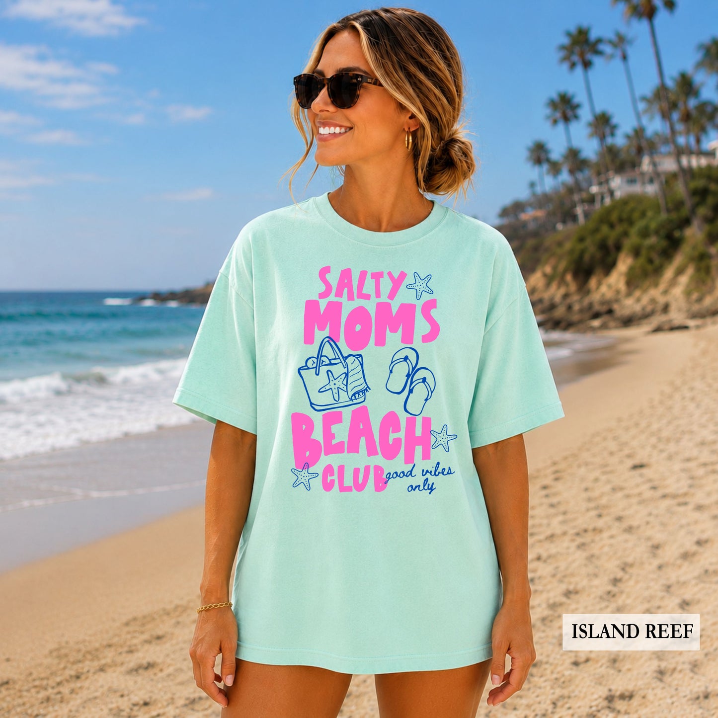 Woman wearing a light blue t-shirt with 'Salty Moms Beach Club' text on a beach, with palm trees and ocean in the background.