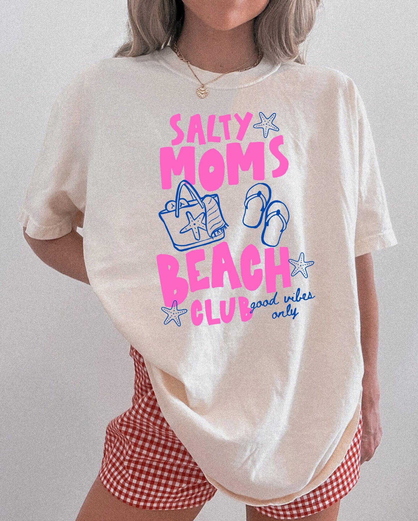 Person wearing a white t-shirt with 'Salty Moms Beach Club' text and graphics on a plain background