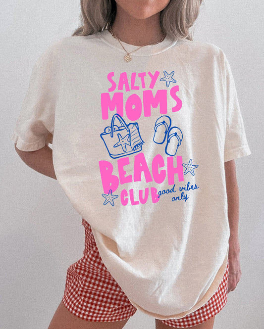 Person wearing a white t-shirt with 'Salty Moms Beach Club' text and graphics on a plain background
