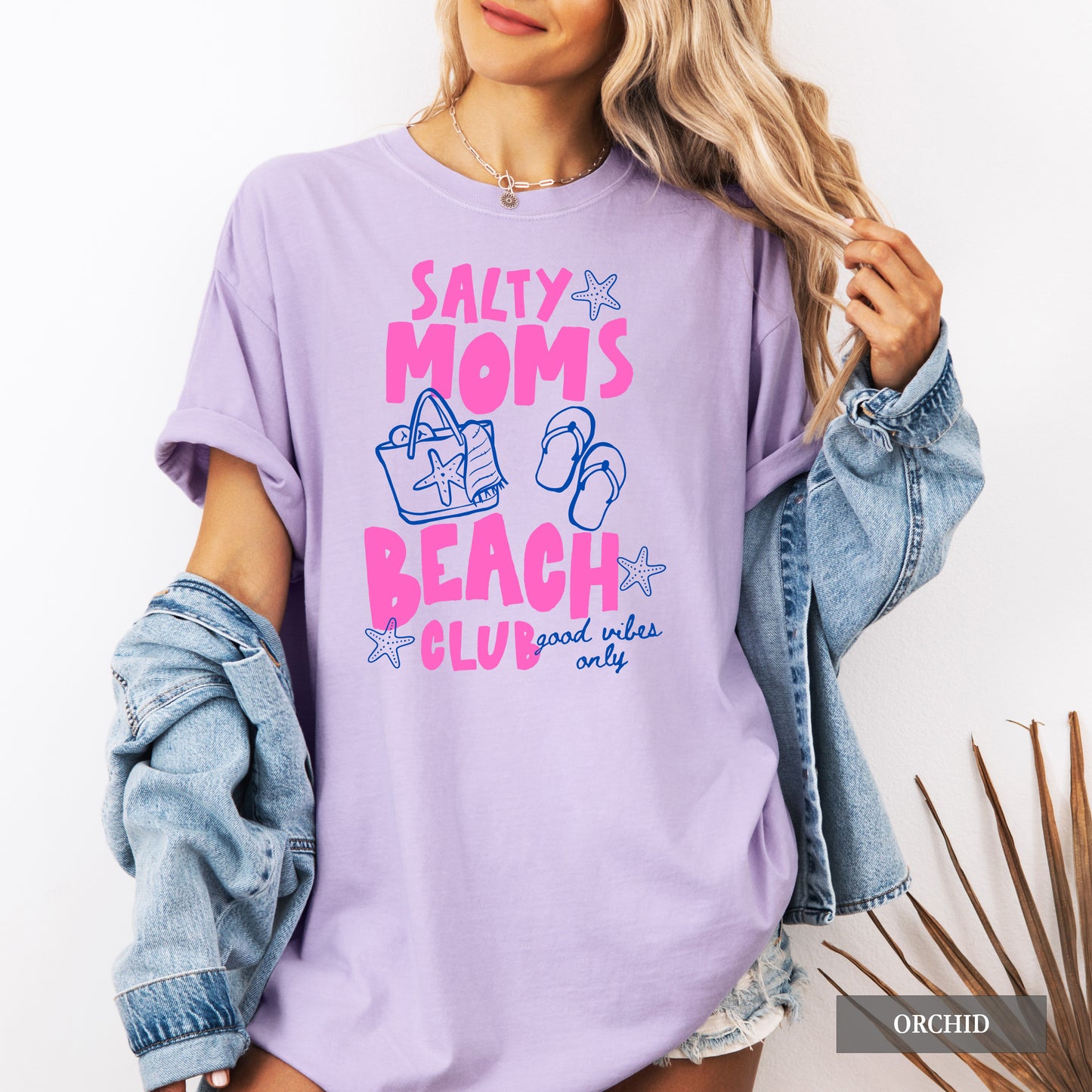 Woman wearing a purple t-shirt with 'Salty Moms Beach Club' text and graphics, holding a denim jacket.