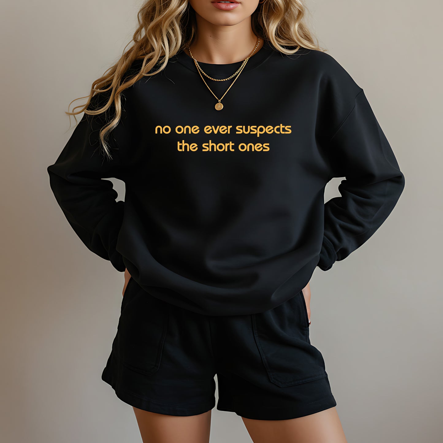 A woman wearing a black crewneck sweatshirt with yellow text that reads 'no one ever suspects the short ones' and matching black shorts, shown against a plain beige background.