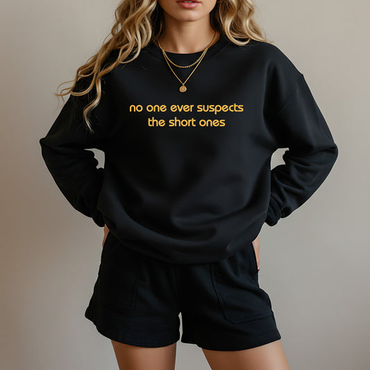 A woman wearing a black crewneck sweatshirt with yellow text that reads 'no one ever suspects the short ones' and matching black shorts, shown against a plain beige background.