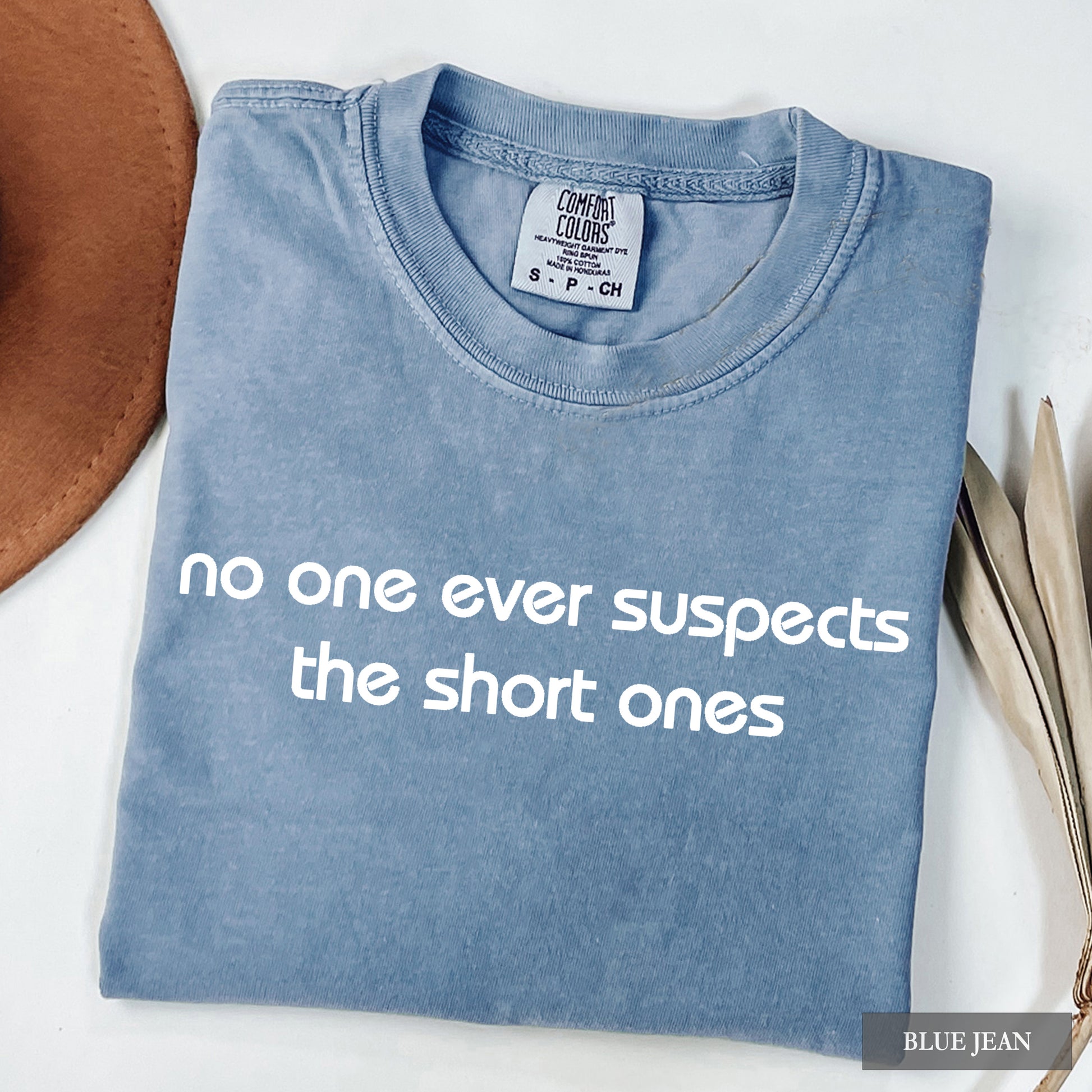 A folded light blue t-shirt with the phrase 'no one ever suspects the short ones' printed in white across the front. The shirt is displayed flat on a white surface with a brown hat and dried leaves as styling props.