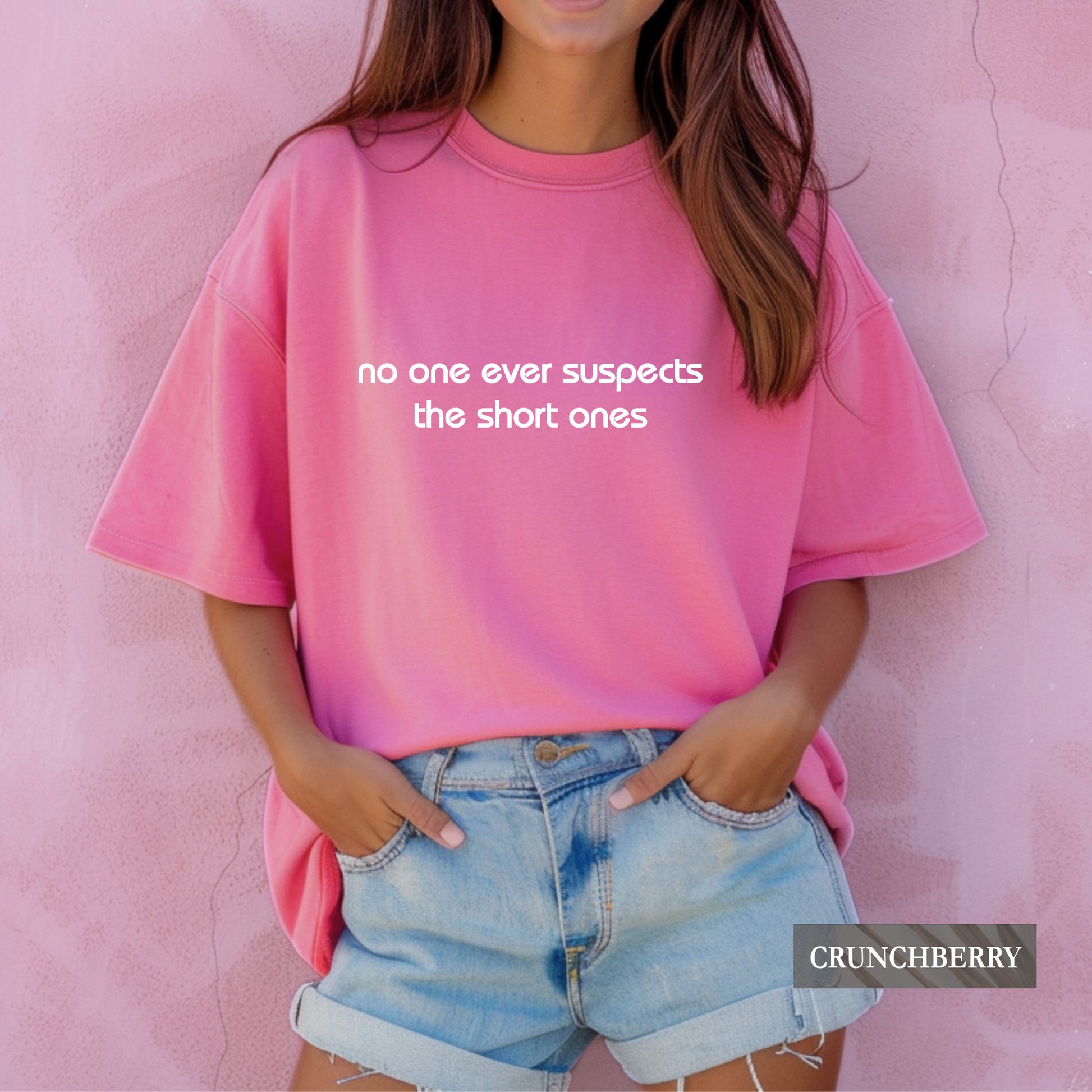 A woman wearing a bright pink oversized t-shirt with the white text 'no one ever suspects the short ones' on the front, paired with light blue denim shorts, standing against a textured pink wall. A 'CRUNCHBERRY' brand label is visible in the bottom right corner.